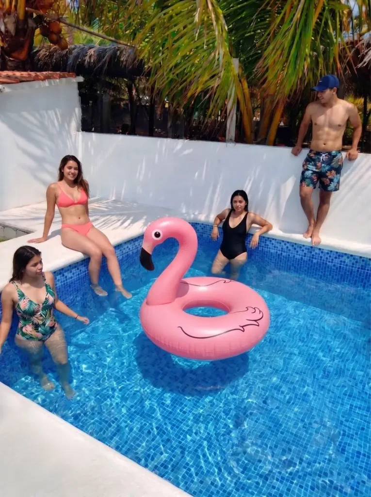 Cuatro jóvenes (tres mujeres y un hombre) disfrutan de un día soleado en la piscina de inmersión de Casa vacacional Don Moy en Chachalacas. Una mujer está de pie en el agua con un traje de baño estampado de palmeras, otra mujer sentada en el borde con un bikini rosa, una tercera mujer de pie en el agua con un traje de baño negro y un hombre de pie en el borde con un bañador estampado y una gorra azul. Un gran inflable de cisne rosa flota en el centro de la piscina azul brillante. Las paredes son blancas y hay una palmera de fondo bajo el sol.